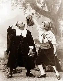 Photo of two white men standing side by side, both amused; the older one is bald and looks up at his wig, caught on a tree; the younger man laughs