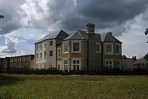 A smartly railed, pale-painted house, with very tall grasses on the main communal garden meadow in front (to south and east), with projecting curved bay and two half-hexagonal bays, and large traditional windows, with a stock-brick three-storey terrace with sash windows and a gable-end fronted house.