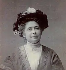 An older white woman, wearing large black hat, a high-collared white lace blouse or dress. Her greying hair is in a bouffant updo.