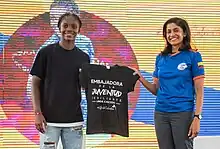 Linda Caicedo holding the left sleeve of a black t-shirt that reads "Embassador of the Resilient Youth."  Anu Rajaraman is to her right, holding the right sleeve of the t-shirt.