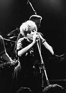 A black-and-white photograph of a woman singing into a microphone. Both her hands are raised and placed on the microphone stand. A man performing bass guitar is visible behind her, as well as drum cymbals and an overhead microphone; another woman's arm and the fretboard of an electric guitar are visible to her right.