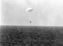 A conical black spacecraft falling towards the surface of the ocean under a single white parachute, seen from some distance away. Very little detail can be seen.