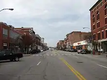 A view down the middle of a city street with two- to four-story brick buildings, and diagonally parked cars, on either side