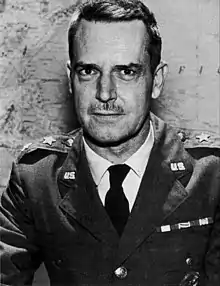 Man with dark hair and moustache in a dress uniform, consisting of a military suit and tie, sitting at a table, with two stars on each lapel indicating his rank, in front of a world map on a wall.