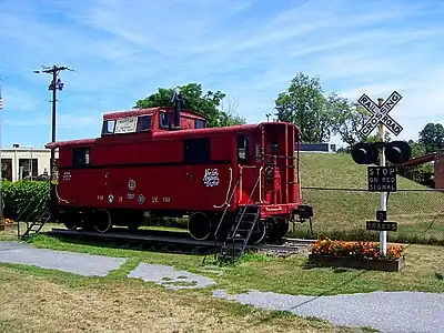 A red caboose on a short set of railroad tracks with a crossing signal at right. Behind them are a chainlink fence and a small rise