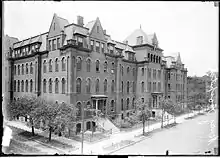 An elevated view of a five-story building on a street corner that takes up a long portion of a block.  The building has multiple entrances leading to the sidewalk on the right.  Trees line the street rising to the height of about two stories.