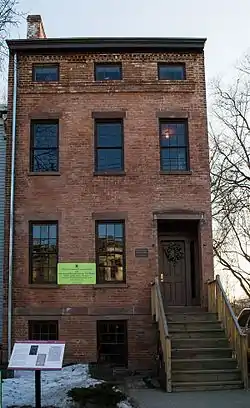 The front of a two-and-a-half story brick building with an exposed basement, seen from a nearby street, partially obstructed by the front of a parked car