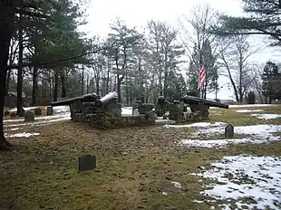 A monument featuring four black cannon barrels mounted on a stone wall in the middle of a small cemetery.  The ground is partly covered with snow.  Many trees stand in the background.  The sky is cloudy.