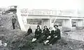 Milton Manaki with fiancée Vasilikija Dauka and other relatives, sitting on riverbank with bridge in background in 1928