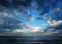 Late daytime view looking far out over an ocean from a beach, which is out of view off the bottom margin. Three-fourths of the shot features a sky marked by heavy cloud cover, which is parting near the middle, revealing a dazzlingly bright cerulean blue sky that darkens near the margins. The ocean is striated with waves coming in parallel to the horizon.