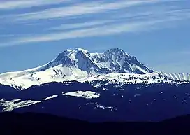 A snow-covered, conical mountain with two peaks rising over the foreground on a nearly clear day