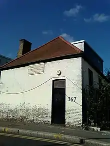 Photograph of a plain white brick building with a red roof and black door