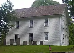 A two-story white building with a pointed roof, seen from the side, with gravestones in front, similarly austere