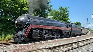 A black streamlined steam locomotive with a 4-8-4 wheel arrangement (four leading wheels, eight driving wheels, and four trailing wheels) and its tender, pulling an observation car with the trees and overhead lines in the background