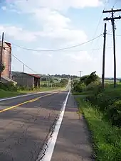 A two-lane highway passes through a rural area. A farm is to the left of the highway; in the background and in the distance are large, open fields.