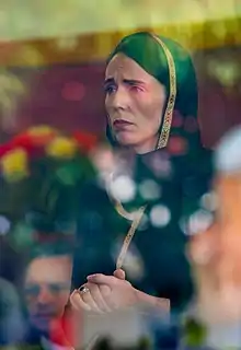 A photo of a woman from the waist up, hands clasped in front of her, with a sad facial expression. She is wearing a black dress and scarf with gold trim.