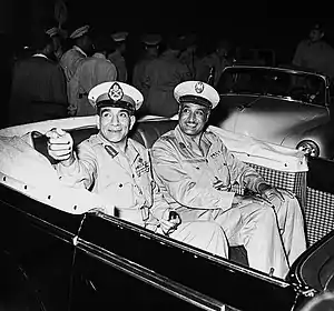 Two smiling men in military uniform seated in an open-top automobile. The first man on the left is pointing his hand in a gesture. Behind the automobile are men in uniform walking away from the vehicle