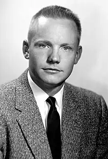 A black-and-white photo of a young man with light skin and pale irises. His mid-colored hair is cut short. He is looking at the camera. He is wearing a barleycorn sport coat, a white shirt and a dark necktie.