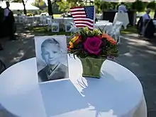 A color image showing a black-and-white photo of a young boy. The picture stands on a small round table beside a vase of flowers containing a U.S. flag.