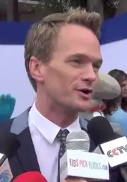 A man with dark blonde hair speaks into microphones away from the camera