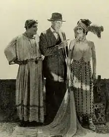 A man in a suit stands between a man and women dressed in costume as figures from the Roman Empire