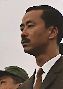 Middle-aged man with side-parted black hair and moustache, in a black suit, white shirt and brown tie. To the left is a clean-shaven Asian man with black hair and a green military cap.