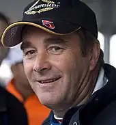 A man in his early fifties is wearing a black baseball cap and is smiling at the camera.