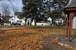 A suburban park, with a large clearing covered in leaves, some trees, and a signpost.