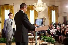 Obama in blue suit standing at a podium in front of an audience as a man in a light grey suit looks on.