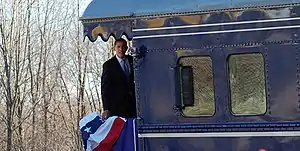 Obama is standing aboard the back platform of a train and looking to the side of the train. There is a red, white, and blue banner hanging over the rear railing.