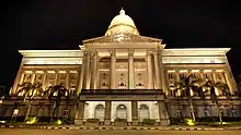 A building with a facade of columns, a dome and a row of palm trees in front, illuminated at night