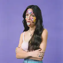 Olivia Rodrigo crossing her arms and sticking her tongue out. Her face is covered in stickers, with the ones on her tongue spelling out "SOUR".