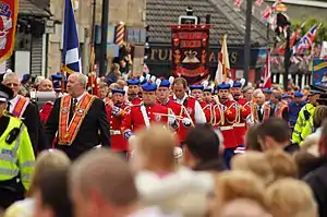 A street is occupied by men and women in formal uniform, some bearing flags, some bearing banners, some playing musical instruments.