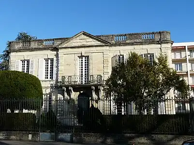 One-storey high building with 5 high windows, with a railing on its flat roof, preceded by a terrace and a fence.