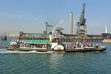 A small, black-hulled paddle steamer passing a commercial wharf, with a power station in the background. The paddle steamer is well loaded with passengers, and has a yellow funnel with black top