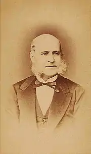 A photograph showing the head and shoulders of an older, balding man with long sideburns dressed in a dark suit, vest and bow tie