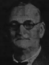 A grainy image of a stern-faced, balding man with glasses.