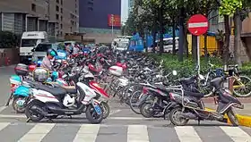 Two rows of motorbikes, many showing their age and use, parked next to a city street corner. There is a large white-bar-on-red-circle "do not enter" sign at the upper right.