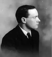 A black and white portrait of a man seated, facing right, wearing a suit with a blank expression.