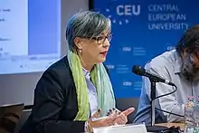 Photograph of a woman wearing glasses, a blue suit and a green scarf sitting in front of a speaker's table with a microphone and a bearded man seated to her left.