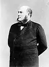 A three-quarters length shot of a Caucasian male with his arms grasping behind his back.  He is wearing dark sideburns, a goatie, black bow tie, white shirt and a black coat.