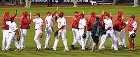 A group of men in white baseball uniforms with red pinstripes and red baseball caps high-five each other while passing in lines moving in opposite directions.