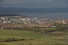 Multiple buildings including terraces, detached houses and blocks of flats. In the foreground are fields and in the background water and then hills.