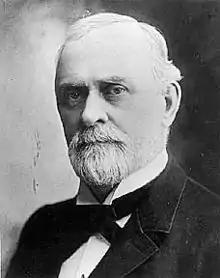 headshot image of a Caucasian male with white hair and a goatie who is wearing a tuxedo with a black tie.