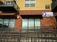 Quatrefoil Library as seen from the sidewalk: the rails of a wheelchair ramp wind up toward a brick building. A sign on the wall reads "Quatrefoil Library". Stenciled on the windows is the abbreviation "Q Library".