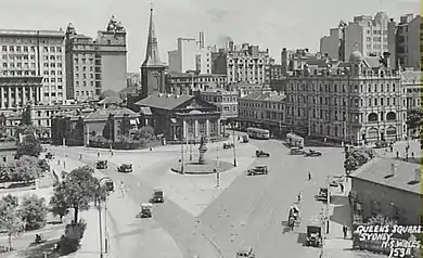 Aerial black and white photo of the junction of Macquarie and King Streets when traffic could still run from one into the other. Buses and other vehicles are on the roads and the now demolished building at the corner can be seen