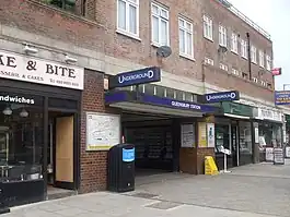 A brown-bricked building with a rectangular, dark blue sign reading "QUEENSBURY STATION" in white letters all under a light blue sky