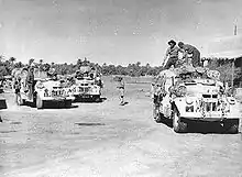 three trucks overloaded with supplies and six men in the foreground. The trees of the oasis can be seen in the background