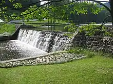 Sunlit water spills over a stone dam in a bright green grassy landscape