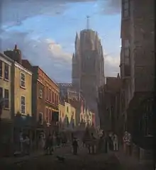 People fill a shadowed street lined with shops and houses in a mixture of architectural styles, leading towards a sunlit church tower